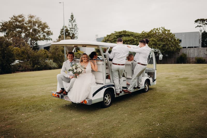 The bride Chloe and groom Brodie sit at the back of a white golf cart at Sandstone Point Hotel, accompanied by wedding party members dressed in light-colored attire, some standing on the side steps of the cart.
