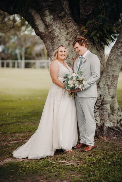 The bride Chloe and groom Brodie stand together under a large tree at Sandstone Point Hotel. Chloe is wearing a white lace wedding gown and holding a bouquet of white and green flowers, while Brodie is dressed in a light gray suit with a white shirt and tie.