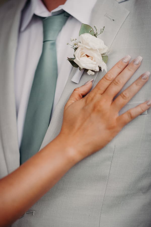 Close-up of the groom's light grey suit jacket with a white floral boutonniere pinned on the lapel and the bride's hand resting on the chest.