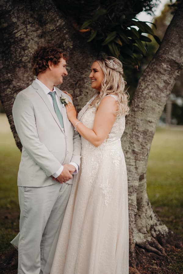 Bride Chloe and groom Brodie stand facing each other in front of a large tree at Sandstone Point Hotel during their couple portraits session. Chloe wears a lace wedding gown with hair accessories, and Brodie wears a light grey suit with a boutonniere and teal tie.
