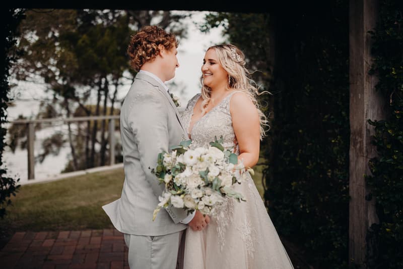 Bride Chloe and groom Brodie stand facing each other holding a bouquet of white flowers at Sandstone Point Hotel outdoors with trees and greenery in the background.