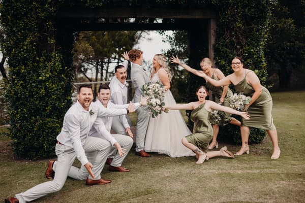 Bride Chloe and groom Brodie stand facing each other under a wooden arch at Sandstone Point Hotel, surrounded by three groomsmen in white shirts and light pants and three bridesmaids in olive green dresses posing playfully around them.