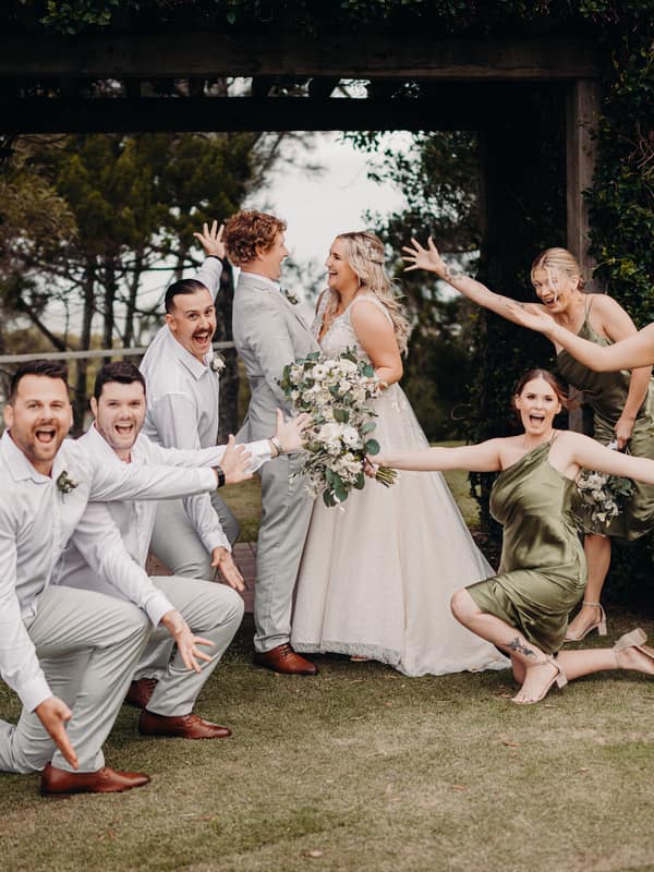 Bride Chloe and groom Brodie stand facing each other under a wooden arch at Sandstone Point Hotel, surrounded by three groomsmen in white shirts and light pants and three bridesmaids in olive green dresses posing playfully around them.