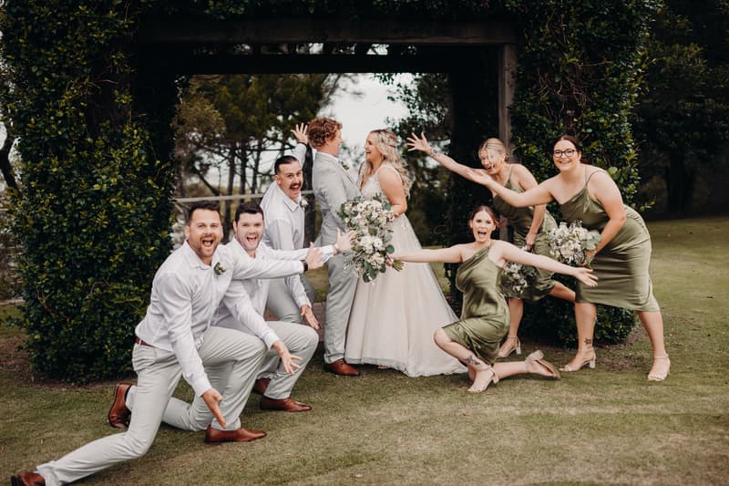 Bride Chloe and groom Brodie stand facing each other under a wooden arch at Sandstone Point Hotel, surrounded by three groomsmen in white shirts and light pants and three bridesmaids in olive green dresses posing playfully around them.