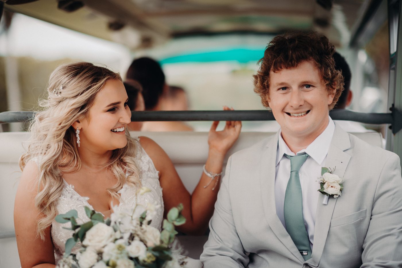 Bride Chloe and groom Brodie sit together inside a vehicle, with Chloe holding a bouquet of white flowers and greenery while smiling at Brodie, who is smiling at the camera wearing a light grey suit with a boutonniere. Candid wedding photographer by She Said Yes.