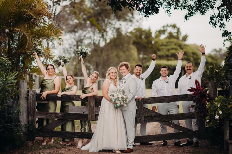 Bride Chloe and groom Brodie stand together in front of a wooden gate at Sandstone Point Hotel, surrounded by three bridesmaids in olive green dresses holding bouquets and three groomsmen in white shirts and light-colored pants, some raising their hands.