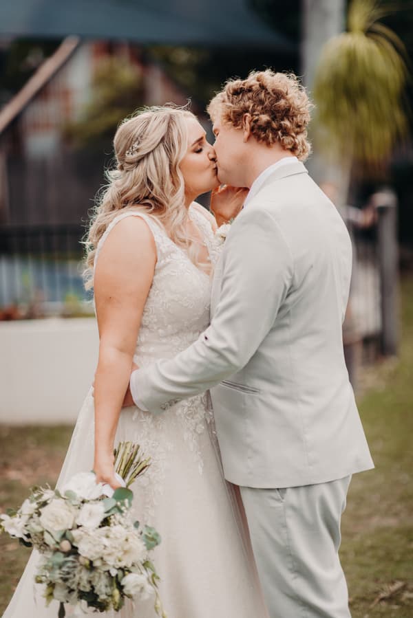 The bride Chloe and groom Brodie share a kiss during their couple portraits at Sandstone Point Hotel. Chloe holds a bouquet of white flowers and wears a lace wedding gown, while Brodie wears a light-colored suit.