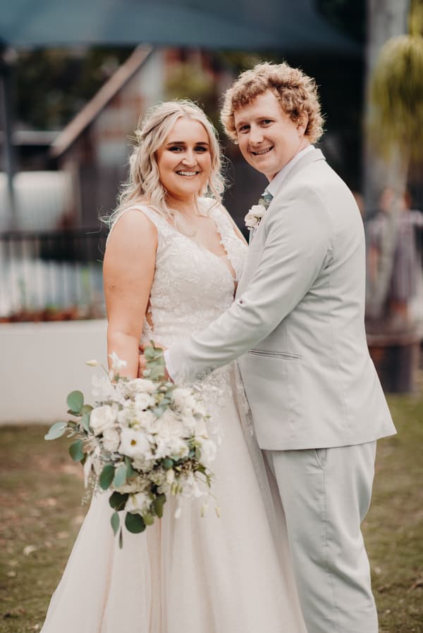 The bride Chloe in a white lace wedding gown holding a bouquet of white flowers and greenery, and the groom Brodie in a light-colored suit, pose together outdoors at Sandstone Point Hotel.