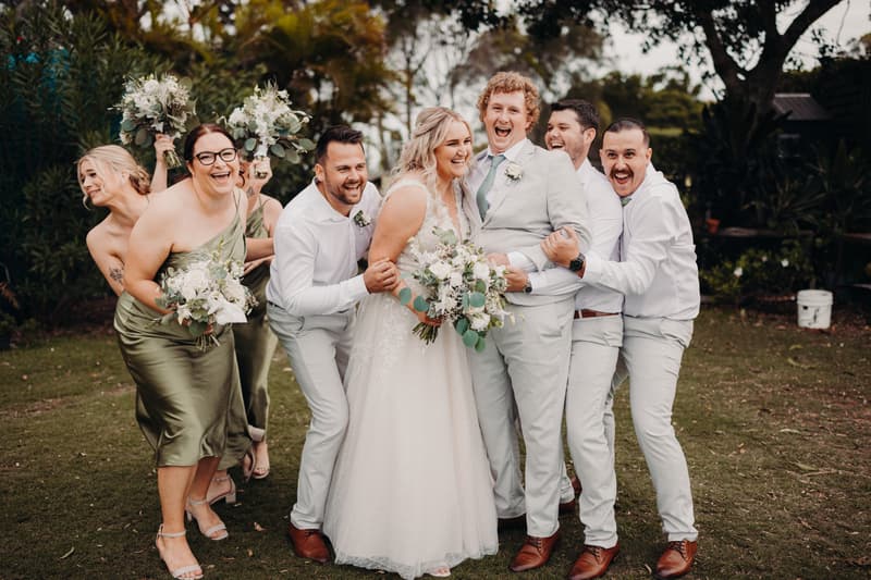 Bride Chloe and groom Brodie pose with bridesmaids and groomsmen outdoors at Sandstone Point Hotel, all smiling and holding bouquets or wearing boutonnieres.