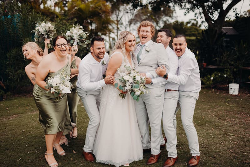 Bride Chloe and groom Brodie pose with bridesmaids and groomsmen outdoors, all smiling and holding bouquets or wearing boutonnieres. Popular wedding seasons 2026 by She Said Yes.