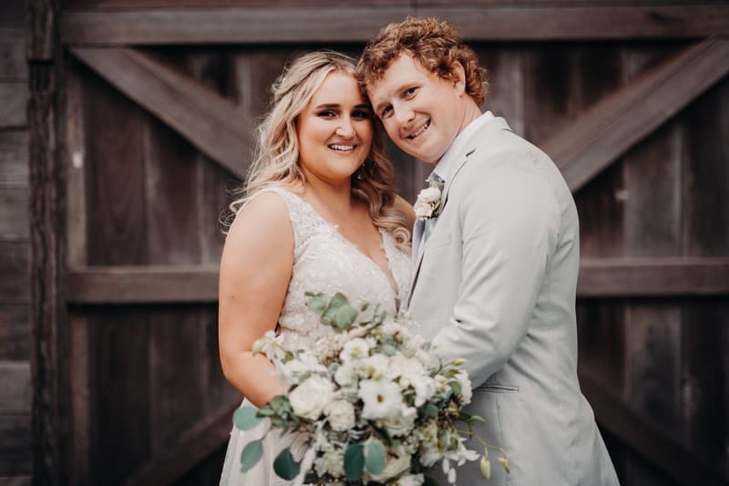 The bride Chloe and groom Brodie pose together holding a bouquet of white flowers in front of a rustic wooden door at Sandstone Point Hotel.