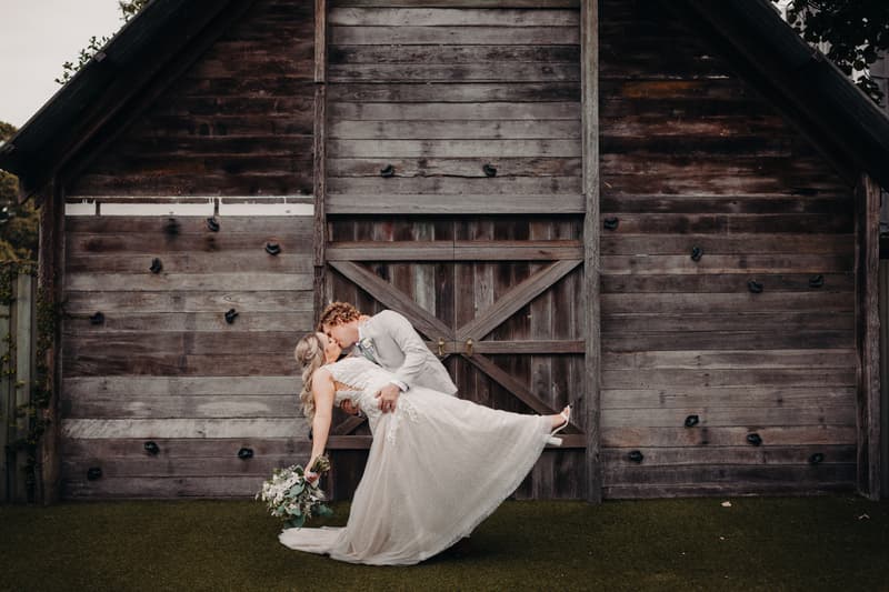 The bride Chloe and groom Brodie share a kiss in front of a large rustic wooden barn door at Sandstone Point Hotel, with Brodie dipping Chloe who holds a bouquet.