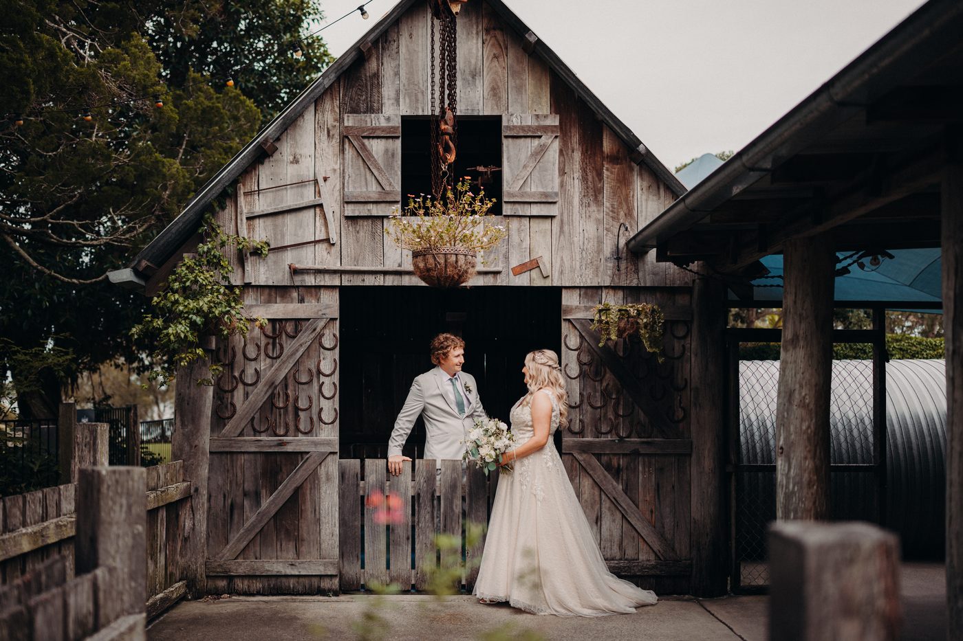 The bride Chloe and groom Brodie stand facing each other in front of a rustic wooden barn door, with the bride holding a bouquet and the groom resting his arm on the half-door. Candid wedding photographer by She Said Yes.