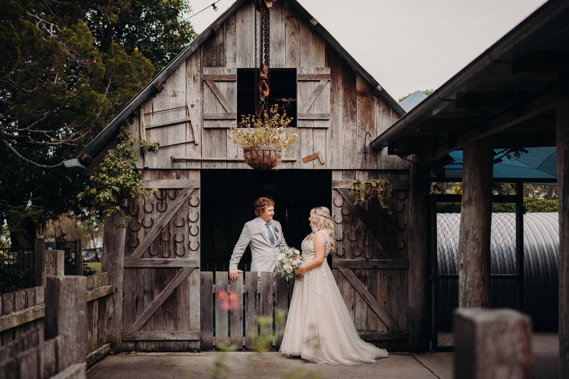 The bride Chloe and groom Brodie stand facing each other in front of a rustic wooden barn door at Sandstone Point Hotel, with the bride holding a bouquet and the groom resting his arm on the half-door.