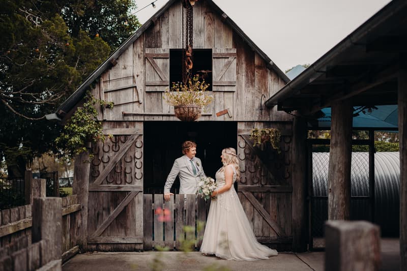 The bride Chloe and groom Brodie stand facing each other in front of a rustic wooden barn door at Sandstone Point Hotel, with the bride holding a bouquet and the groom resting his arm on the half-door.