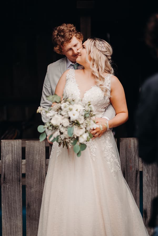 Bride Chloe in a white lace wedding gown holding a bouquet of white flowers and greenery, embraced from behind by groom Brodie in a light grey suit, posing in front of a wooden fence at Sandstone Point Hotel.