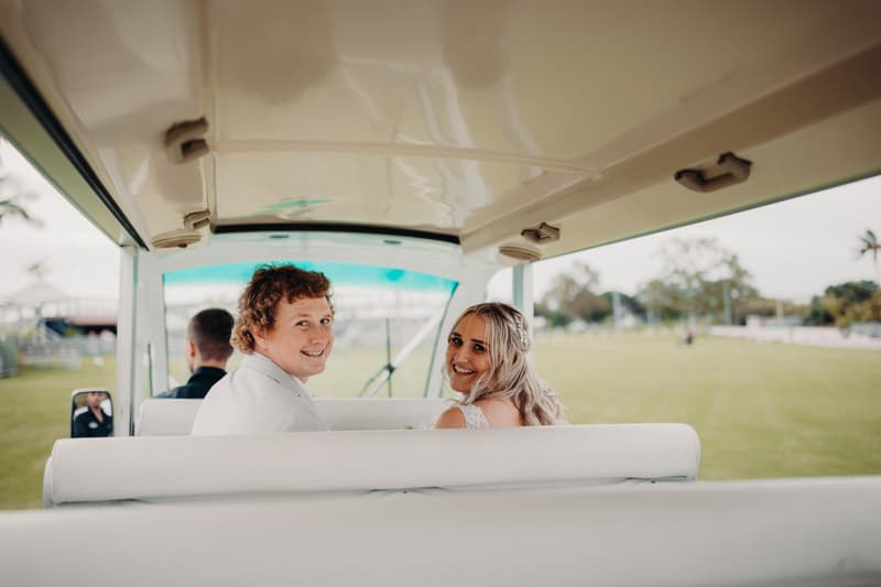The bride Chloe and groom Brodie sit together in a white golf cart at Sandstone Point Hotel, both looking back and smiling towards the camera with a grassy field and trees visible outside.