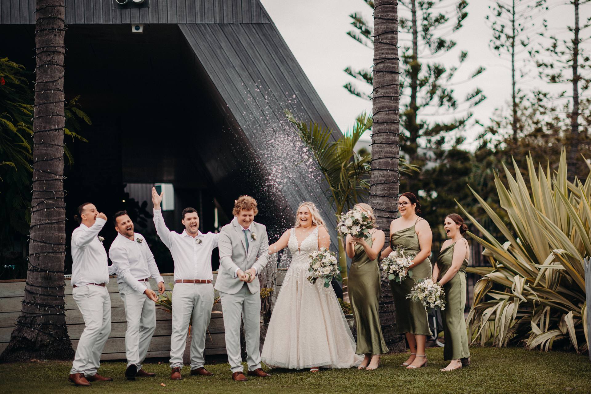 The bride Chloe and groom Brodie stand together at Sandstone Point Hotel with their wedding party. Brodie is spraying champagne while the bridesmaids in olive green dresses and groomsmen in white shirts and light pants react with joy and laughter outdoors near palm trees and greenery.