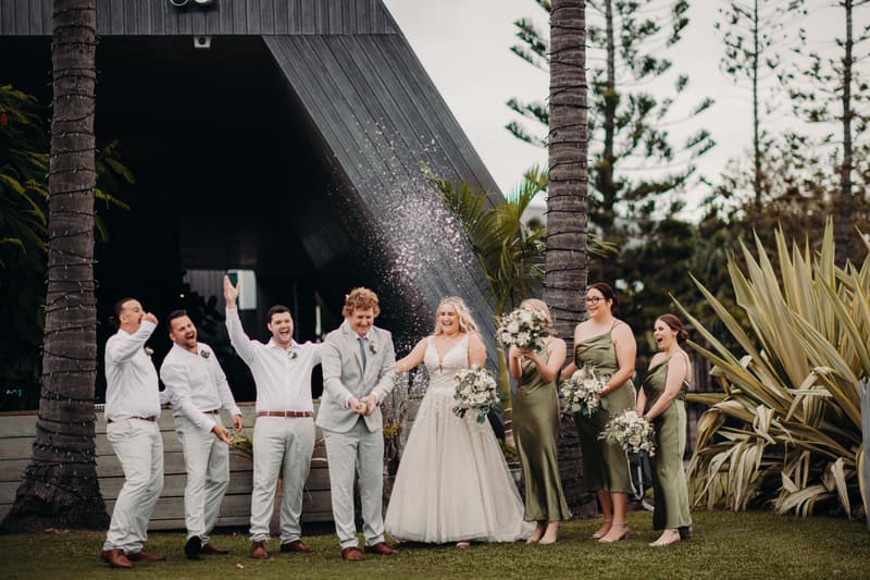 The bride Chloe and groom Brodie stand together at Sandstone Point Hotel with their wedding party. Brodie is spraying champagne while the bridesmaids in olive green dresses and groomsmen in white shirts and light pants react with joy and laughter outdoors near palm trees and greenery.
