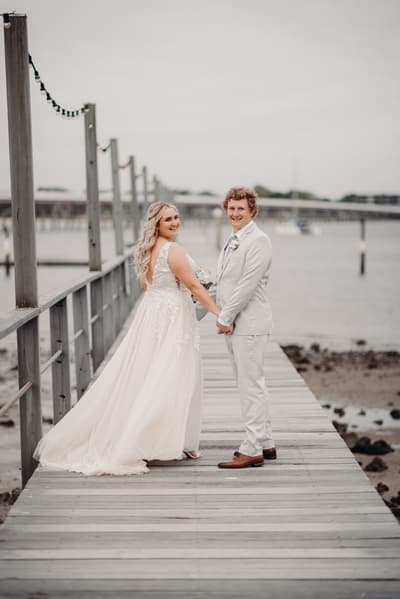 Bride Chloe and groom Brodie hold hands and smile while standing on a wooden pier, with water and a distant bridge in the background. Photo booth hire Sunshine Coast by She Said Yes.