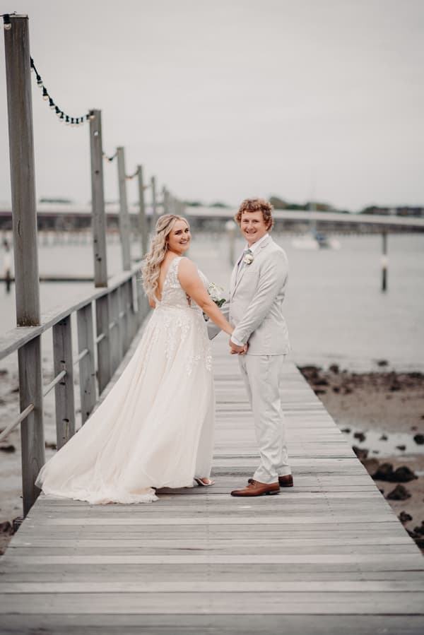 Bride Chloe and groom Brodie hold hands and smile while standing on a wooden pier at Sandstone Point Hotel, with water and a distant bridge in the background.