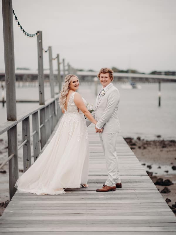 Bride Chloe and groom Brodie hold hands and smile while standing on a wooden pier at Sandstone Point Hotel, with water and a distant bridge in the background.