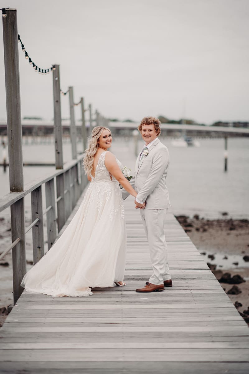 Bride Chloe and groom Brodie hold hands and smile while standing on a wooden pier, with water and a distant bridge in the background. Cinematic wedding photos by She Said Yes.