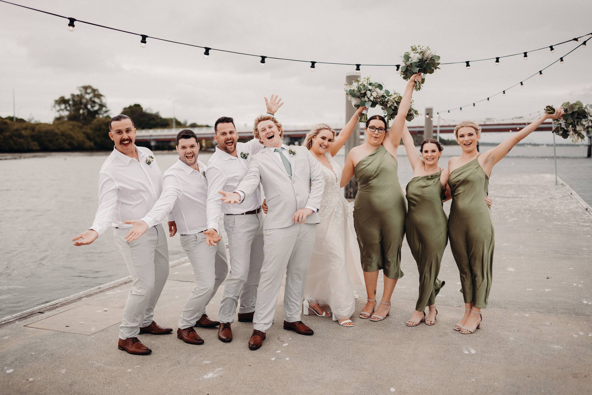 Brodie in a light grey suit and Chloe in a white wedding dress pose with their bridal party on a pier at Sandstone Point Hotel, with bridesmaids in olive green dresses holding bouquets and groomsmen in white shirts and light grey pants.
