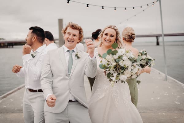 Brodie in a light suit and Chloe in a white wedding gown holding a bouquet pose with two bridesmaids and groomsmen on a pier. Outdoor photo booth hire by She Said Yes.