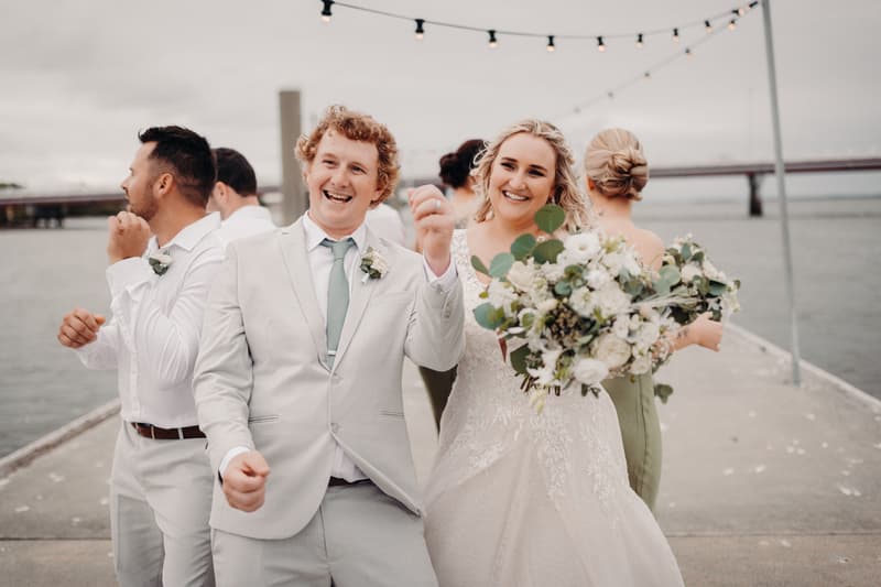 Brodie in a light suit and Chloe in a white wedding gown holding a bouquet pose with two bridesmaids and groomsmen on a pier at Sandstone Point Hotel with water and a bridge in the background.