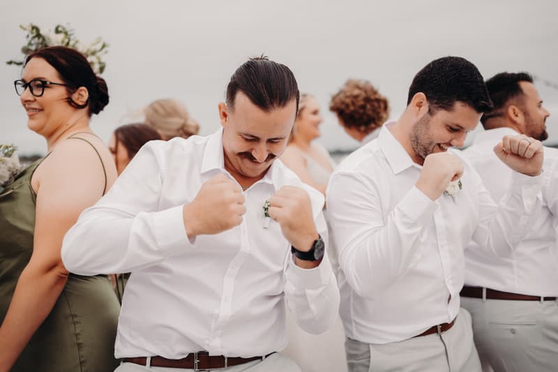 Wedding party members in white shirts and light-colored pants pose together outdoors at Sandstone Point Hotel, with two men in the foreground raising their fists and smiling.