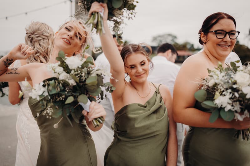 Three bridesmaids in green dresses hold bouquets and smile outdoors at Sandstone Point Hotel, with the bride partially visible in the background.