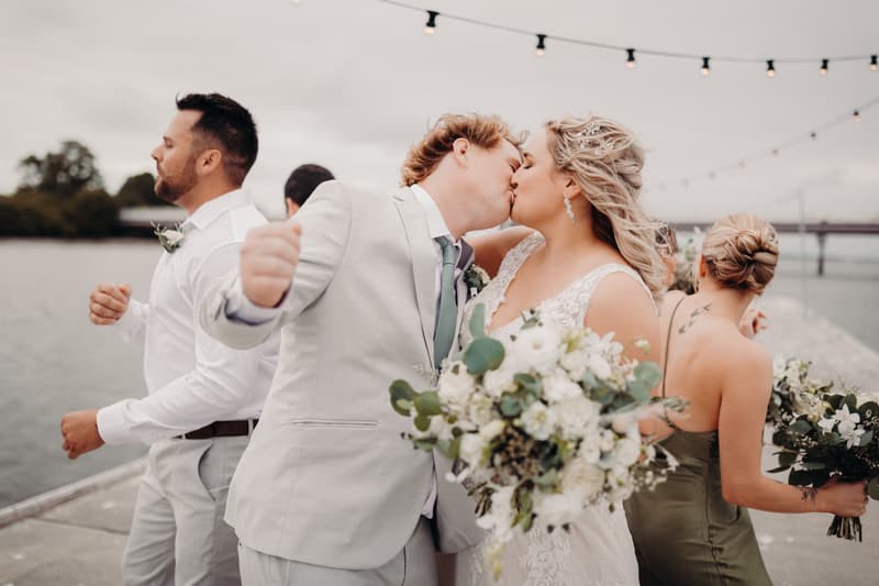 Bride Chloe and groom Brodie kiss while standing on a waterfront pier at Sandstone Point Hotel, accompanied by a bridesmaid in a green dress and a groomsman in a white shirt and light pants.