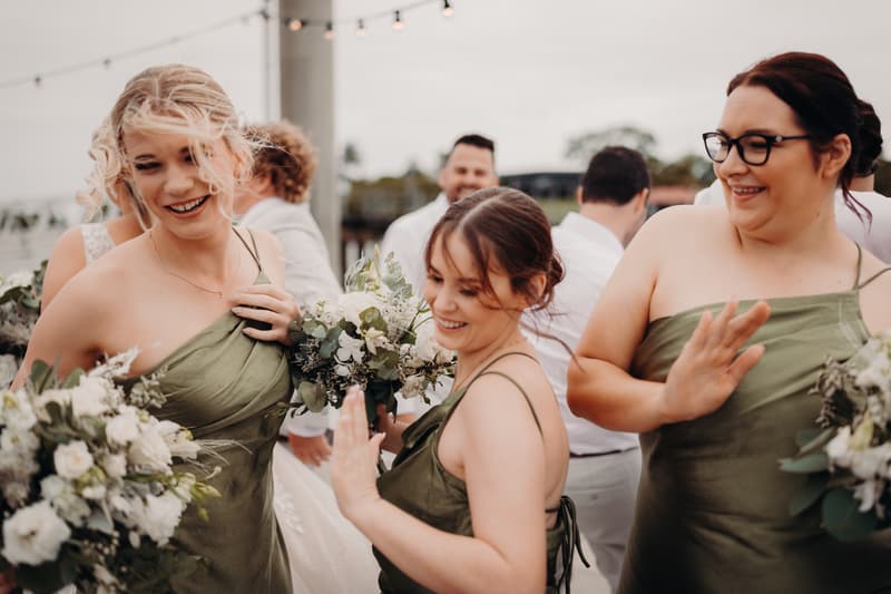 Three bridesmaids in olive green dresses holding bouquets and smiling at Sandstone Point Hotel, with guests blurred in the background.