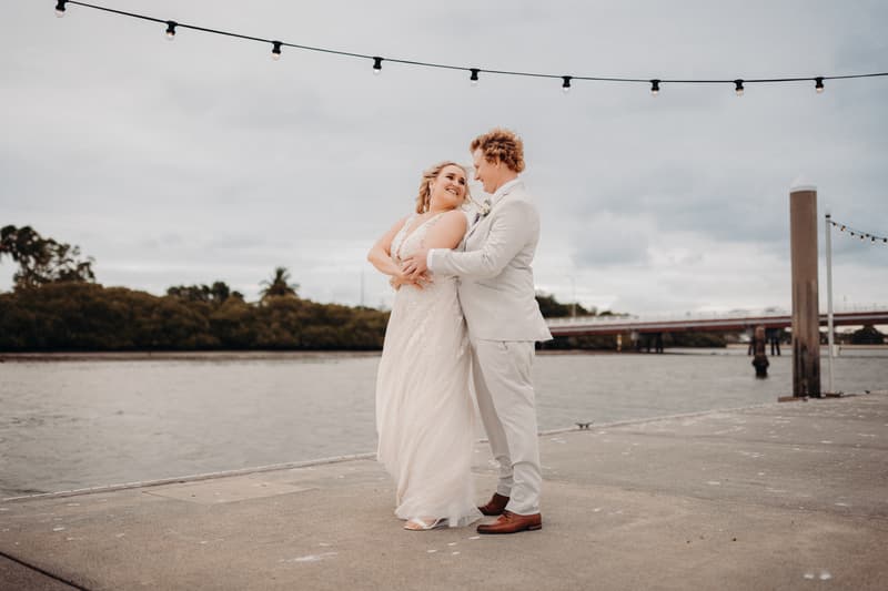 The bride Chloe and groom Brodie embrace on a concrete pier by the water at Sandstone Point Hotel, with string lights overhead and a bridge in the background.
