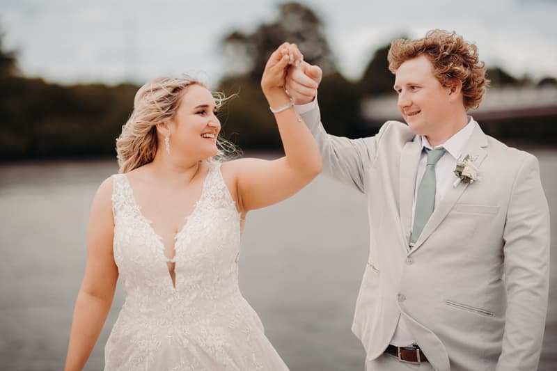 The bride Chloe and groom Brodie hold hands and smile at each other during their couple portraits at Sandstone Point Hotel, with water and trees in the background.
