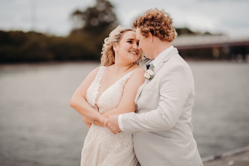 The bride Chloe and groom Brodie embrace and smile at each other near the water at Sandstone Point Hotel.