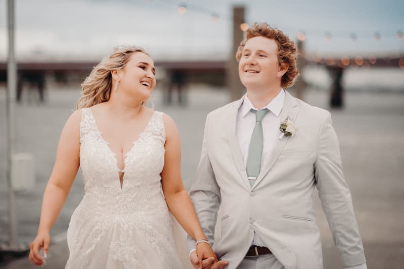 The bride Chloe and groom Brodie walk hand in hand outdoors near a waterfront at Sandstone Point Hotel, both smiling and dressed in wedding attire.