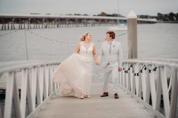The bride Chloe and groom Brodie walk hand in hand on a wooden jetty, with water and a distant bridge in the background. ABIA Awards wedding photography by She Said Yes.