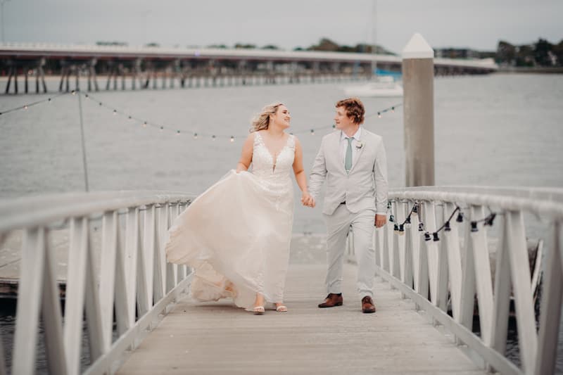 The bride Chloe and groom Brodie walk hand in hand on a wooden jetty at Sandstone Point Hotel, with water and a distant bridge in the background.