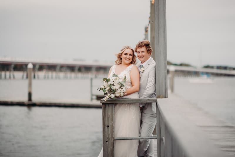 The bride Chloe and groom Brodie pose together on a wooden pier at Sandstone Point Hotel, with water and a distant bridge in the background. Chloe holds a bouquet of white flowers while Brodie stands close behind her.