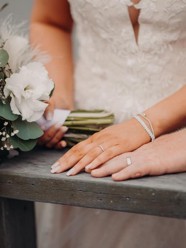 The bride Chloe and groom Brodie rest their hands on a wooden surface at Sandstone Point Hotel, with Chloe holding a bouquet of white flowers and greenery.