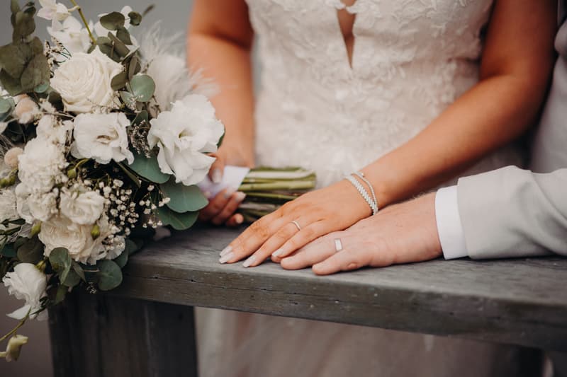 The bride Chloe and groom Brodie rest their hands on a wooden surface at Sandstone Point Hotel, with Chloe holding a bouquet of white flowers and greenery.