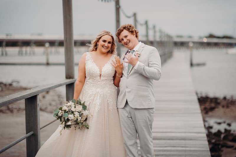 The bride Chloe and groom Brodie stand together on a wooden pier at Sandstone Point Hotel, both showing their wedding rings. Chloe holds a bouquet of white and green flowers and wears a lace wedding gown, while Brodie wears a light grey suit with a boutonniere.