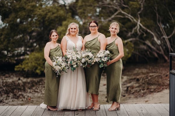 The bride Chloe stands with three bridesmaids dressed in olive green dresses holding bouquets. Photo and video services by She Said Yes.