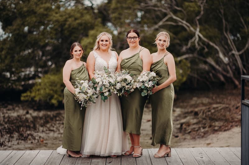 The bride Chloe stands with three bridesmaids dressed in olive green dresses holding bouquets at Sandstone Point Hotel outdoors on a wooden deck with trees in the background.