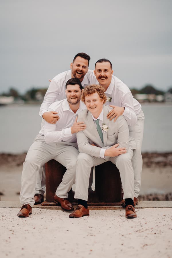 Brodie in a light grey suit with a boutonniere sits on a wooden block at Sandstone Point Hotel, surrounded closely by three groomsmen in white shirts and light-colored pants, all smiling.
