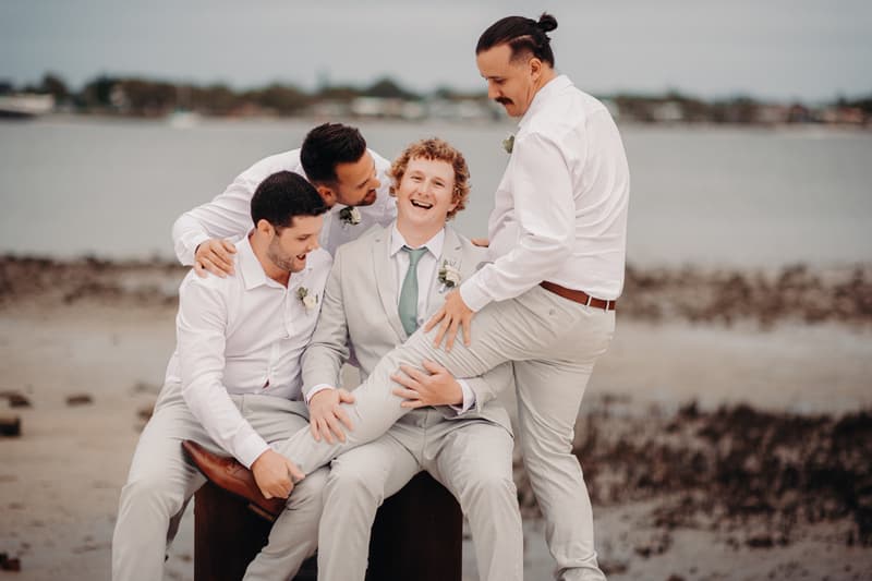 Brodie in a light grey suit with a green tie sits on a wooden block at Sandstone Point Hotel beach while three groomsmen in white shirts and light trousers playfully hold his leg.
