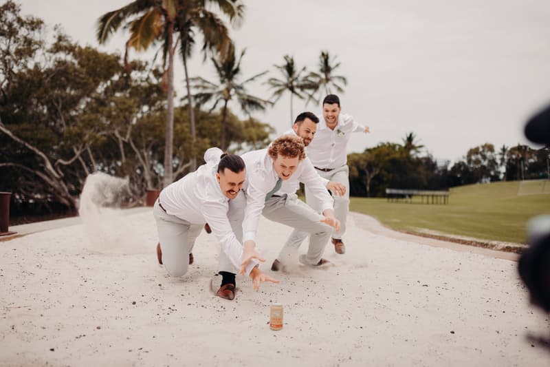 Four groomsmen dressed in white shirts and light-colored pants are playfully lunging forward on a sandy area at Sandstone Point Hotel, kicking up sand behind them.