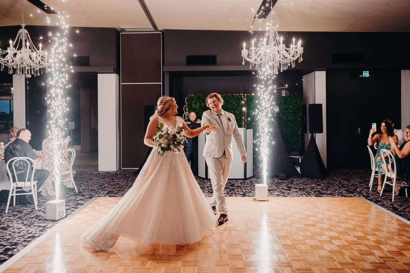 The bride Chloe and groom Brodie dance on the dance floor at Sandstone Point Hotel — Pumicestone Room, surrounded by guests seated at tables and indoor chandeliers, with sparklers creating a celebratory atmosphere.